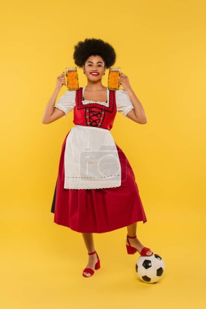 cheerful african american waitress in dirndl standing with beer mugs near soccer ball on yellow