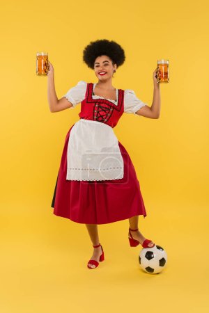 fröhliche afrikanisch-amerikanische Kellnerin im Dirndl mit Bierkrügen in der Nähe des Fußballs auf gelb