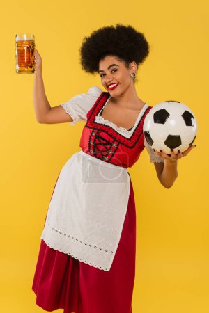 Unbekümmerte afrikanisch-amerikanische Oktoberfestkellnerin im Dirndl mit Bierkrug und Fußball auf gelb