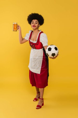 pleased african american waitress in bavarian costume with beer mug and soccer ball on yellow