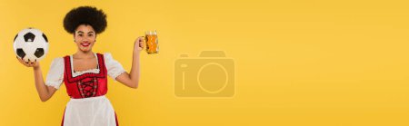 happy african american bavarian waitress in dirndl with beer mug and soccer ball on yellow, banner
