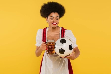 fröhliche afrikanisch-amerikanische Oktoberfest-Kellnerin im Dirndl klingelt Bierkrug mit Fußball auf gelb