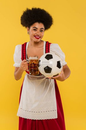 fröhliche afrikanisch-amerikanische Oktoberfest-Kellnerin klimpert mit Bierkrug und Fußball auf gelb