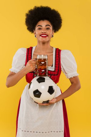 happy african american bavarian waitress with beer mug and soccer ball smiling on yellow