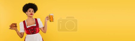 african american bavarian waitress in dirndl holding beer mug and tasty pretzels on yellow, banner