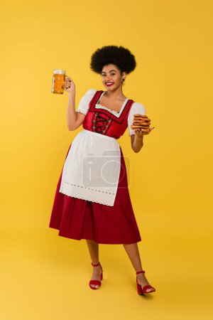 cheerful african american bavarian waitress in dirndl holding beer mug and tasty pretzels on yellow