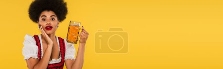 étonnante serveuse afro-américaine oktoberfest avec tasse de bière regardant la caméra sur jaune, bannière