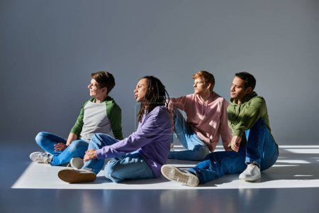 Foto de Cuatro hombres jóvenes en trajes casuales sentados en el suelo sobre fondo gris y mirando hacia otro lado, diversidad - Imagen libre de derechos