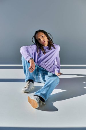 Photo for Handsome african american man in sweatshirt with dreadlocks sitting on floor and looking at camera - Royalty Free Image