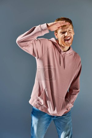 fröhlicher junger Mann in trendigem Kapuzenpulli und Jeans, die Hand an der Stirn, posierend vor grauem Hintergrund