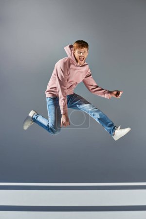cheerful young man in casual clothing jumping and playing invisible guitar on grey backdrop
