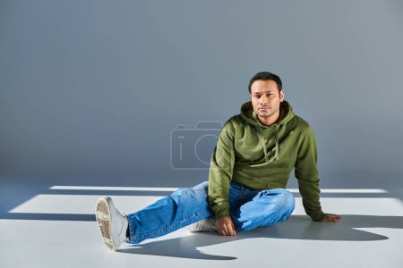 Foto de Guapo joven indio hombre en sudadera con capucha caqui y jeans sentado en el suelo y mirando a la cámara - Imagen libre de derechos