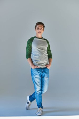 Téléchargez les photos : Jeune homme souriant en casual manches longues et jeans souriant à la caméra posant avec les mains dans les poches - en image libre de droit