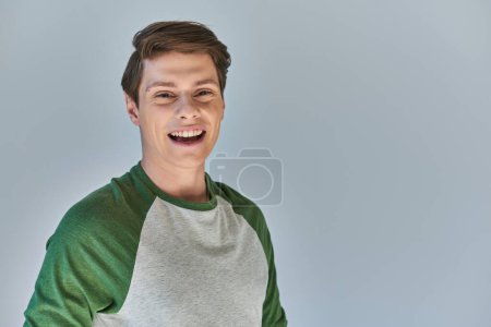 Porträt eines lachenden jungen Mannes in lässiger Stadtkleidung, der vor grauem Hintergrund in die Kamera blickt
