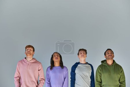 Foto de Cuatro jóvenes sonrientes con atuendo informal brillante mirando hacia arriba en el fondo gris, diversidad cultural - Imagen libre de derechos