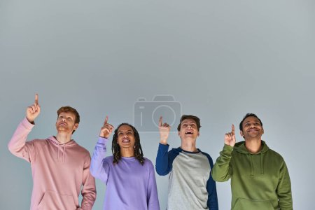 Foto de Cuatro hombres alegres en ropa de calle mirando hacia arriba y señalando hacia arriba sobre fondo gris, diversidad cultural - Imagen libre de derechos