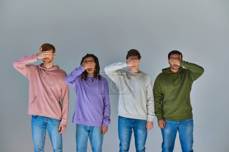 czterech wielorasowych przyjaciół w luźnych ubraniach ulicznych przykrywających oczy dłońmi na szarym tle, różnorodność