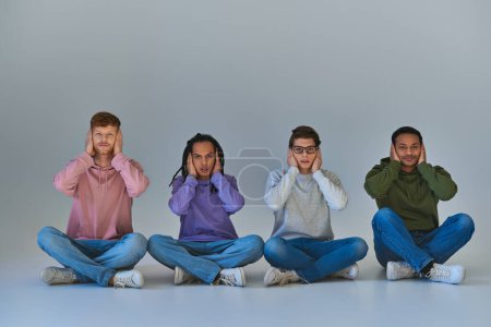 Foto de Cuatro amigos en ropa elegante sentado con las piernas cruzadas y cubriendo las orejas con las manos, diversidad - Imagen libre de derechos