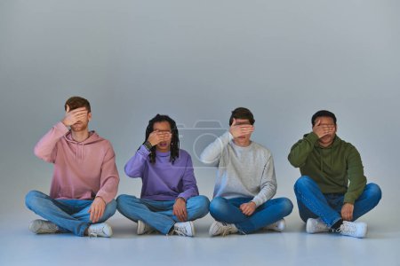 wielokulturowi mężczyźni w modnych strojach siedzący ze skrzyżowanymi nogami zakrywający oczy dłońmi, różnorodność