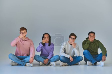 Foto de Cuatro amigos multiculturales sentados con las piernas cruzadas y cubriendo bocas, diversidad cultural - Imagen libre de derechos