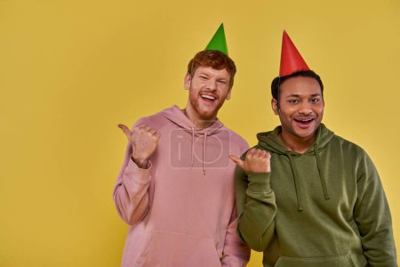 Foto de Dos jóvenes amigos en atuendo casual y sombreros de cumpleaños sonriendo a la cámara señalando los pulgares a un lado - Imagen libre de derechos