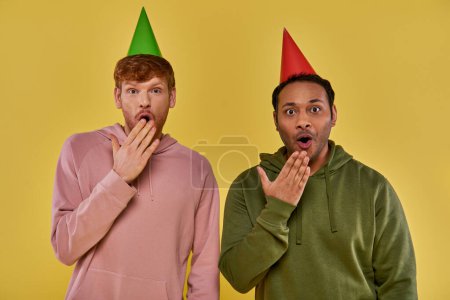 Photo for Astonished multicultural men in everyday street wear in birthday hats covering mouths with hands - Royalty Free Image