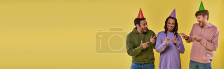 Foto de Tres hombres multiétnicos en sombreros de cumpleaños sonriendo y señalando con los dedos a la cámara, cumpleaños, pancarta - Imagen libre de derechos