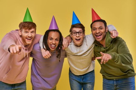 Foto de Amigos multiculturales en sombreros de cumpleaños abrazándose mirando y sonriendo a la cámara, cumpleaños - Imagen libre de derechos