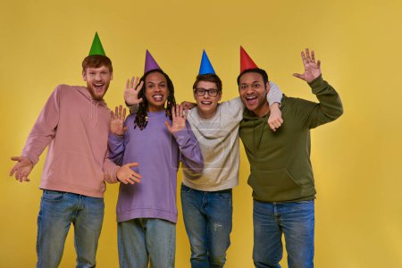 Foto de Cuatro jóvenes amigos en sombreros de cumpleaños sonriendo a la cámara saludando y mostrando las palmas abiertas, cumpleaños - Imagen libre de derechos