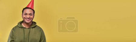 lächelnder indischer Mann in khakifarbenem Kapuzenpulli und Geburtstagsmütze mit Blick in die Kamera auf gelbem Hintergrund, Geburtstag