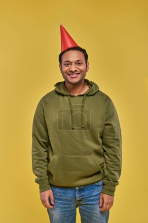 Téléchargez les photos : Joyeux homme indien en tenue décontractée et chapeau d'anniversaire souriant à la caméra sur fond jaune, anniversaire - en image libre de droit
