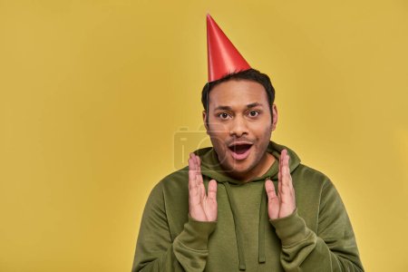 Foto de Asombrado joven indio en sombrero de cumpleaños y sudadera con capucha caqui con las manos más cerca de la cara, cumpleaños - Imagen libre de derechos