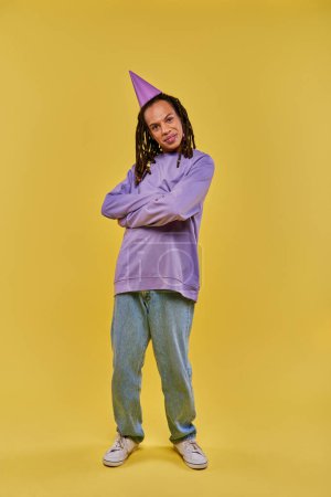 Téléchargez les photos : Homme afro-américain souriant en sweat-shirt violet et chapeau d'anniversaire posant avec les bras croisés sur la poitrine - en image libre de droit