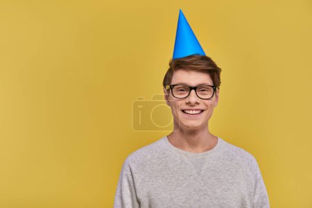 young cheerful man in white sweatshirt and birthday hat smiling at camera on yellow backdrop