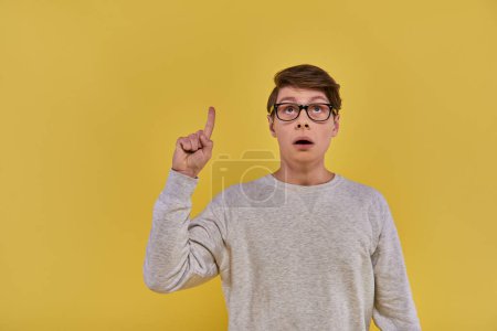 Photo for Young man in white sweatshirt raising his finger in eureka gesture and looking up on yellow backdrop - Royalty Free Image