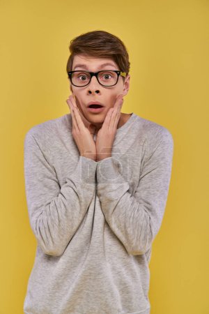 schockierter junger Mann in Freizeitkleidung mit Brille, die seine Wangen mit den Händen auf gelbem Hintergrund berührt