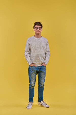 junger gutaussehender Mann in Sweatshirt und Jeans, der still steht und die Hände in den Taschen in die Kamera blickt