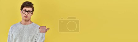 Photo for Young man in simple wear and glasses pointing finger aside on left side on yellow backdrop, banner - Royalty Free Image