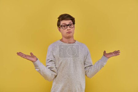 zdezorientowany młody człowiek w bluzie i okularach pokazujący bezradny gest na żółtym tle
