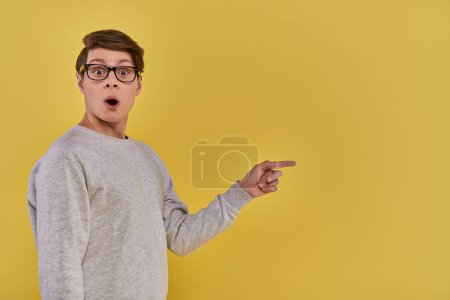 Téléchargez les photos : Homme joyeux étonné en tenue décontractée avec des lunettes pointant du doigt sur le côté droit, fond jaune - en image libre de droit
