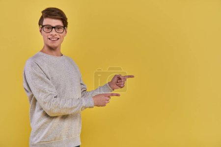 Téléchargez les photos : Joyeux jeune homme en tenue décontractée pointant deux doigts de côté sur le côté droit sur fond jaune - en image libre de droit