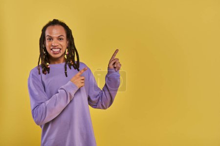 Téléchargez les photos : Joyeux homme afro-américain avec dreadlocks percé lèvre pointant sur le côté droit sur fond jaune - en image libre de droit