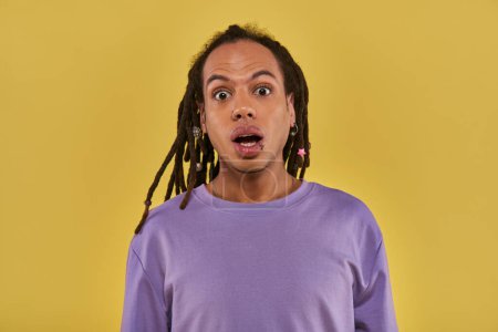 Téléchargez les photos : Gros plan surpris homme en sweat-shirt violet avec lèvre percée et dreadlocks sur fond jaune - en image libre de droit