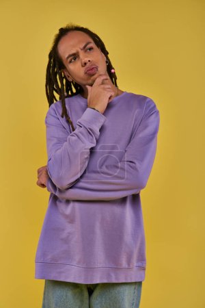 Téléchargez les photos : Réfléchi jeune homme en sweat-shirt violet penser et regarder vers le haut et loin sur fond jaune - en image libre de droit