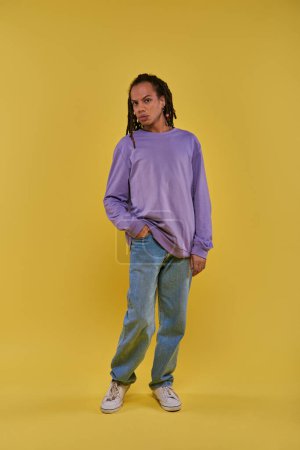 Téléchargez les photos : Jeune afro-américain en sweat-shirt violet et jeans avec dreadlocks posant sur fond jaune - en image libre de droit