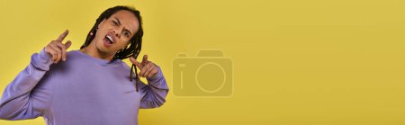 Foto de Desconcertado y sorprendido hombre afroamericano en sudadera púrpura con dedos punzantes piercing, pancarta - Imagen libre de derechos