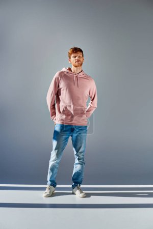 handsome red haired man in pink hoodie posing with hands in pockets of jeans on grey backdrop
