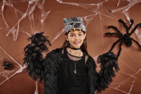cheerful preteen girl posing in black Halloween outfit and wolf mask on brown background