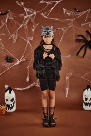 linda chica en traje de piel sintética con máscara de lobo de pie todavía sobre fondo marrón, concepto de Halloween