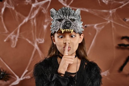 Nahaufnahme eines vorpubertierenden Mädchens in Wolfsmaske, das Schweigen zeigt und Finger betrachtet, Halloween-Konzept